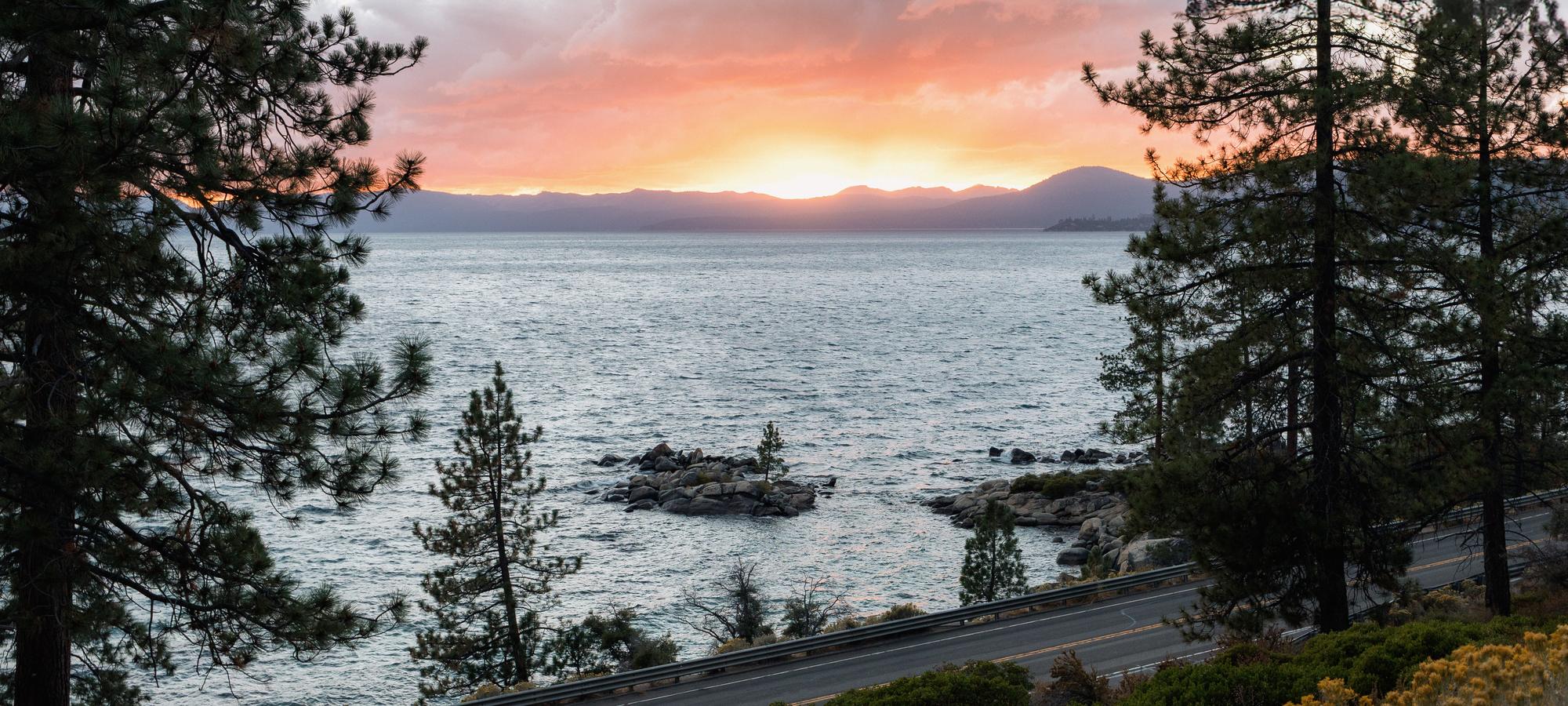 Highway 28 along Lake Tahoe's East Shore at sunset, pines on both sides and granite boulders in the water.