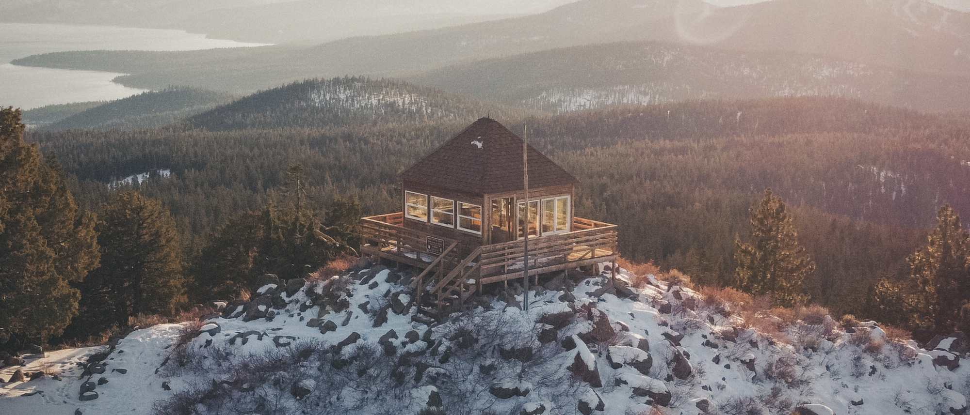 The Martis Peak fire lookout on a snowy, rocky summit above miles of pine forest, a lake far off in the haze.
