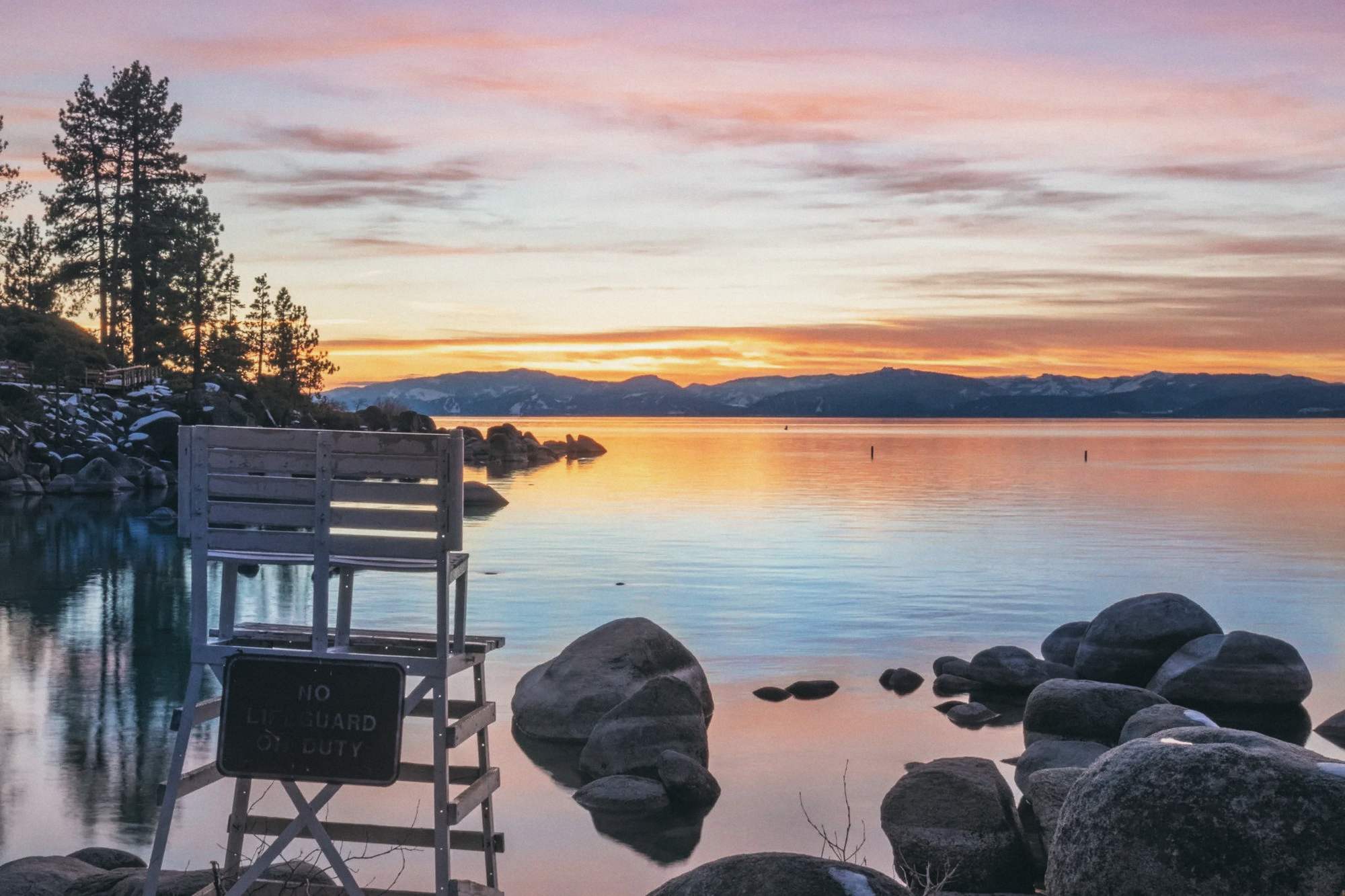 Sunset across Lake Tahoe, with granite boulders and a lifeguard chair on the shore.