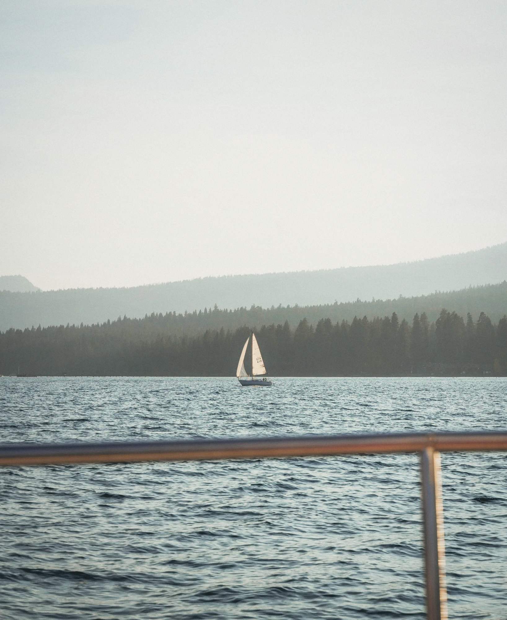 A lone white sailboat on open water at Lake Tahoe, seen past a brass deck rail.