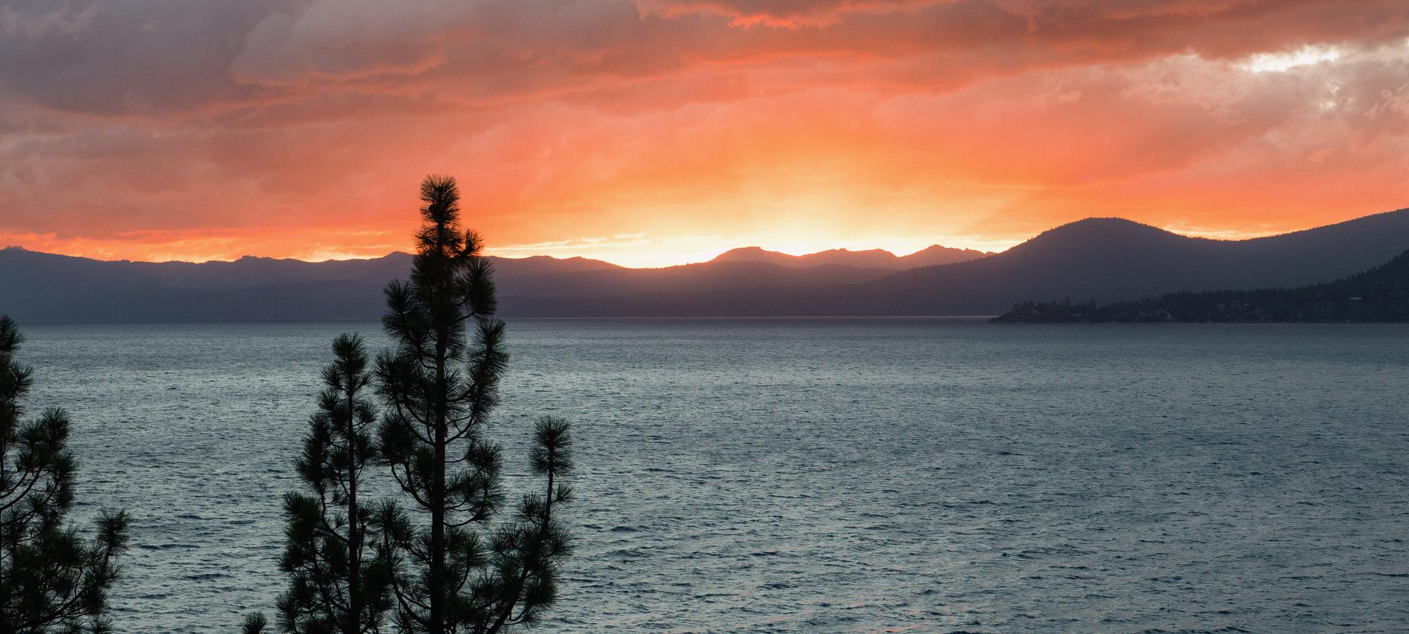 Sunset over Lake Tahoe through pines on the East Shore, the far shore's mountains in silhouette.