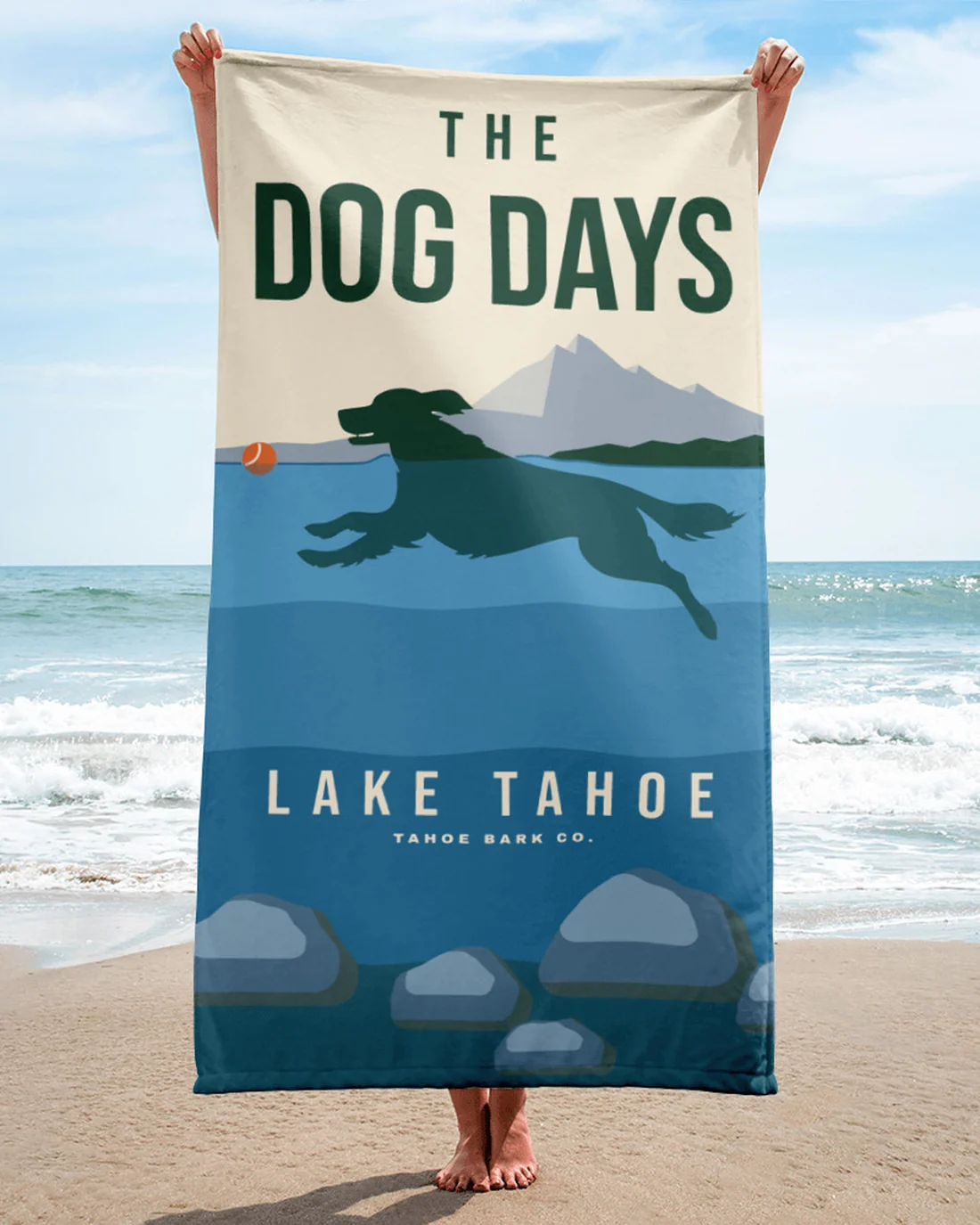 The Dog Days beach towel held up on a sandy beach, the whole print showing: THE DOG DAYS over the peak, the dog and the ball at the waterline, LAKE TAHOE in the blue water.