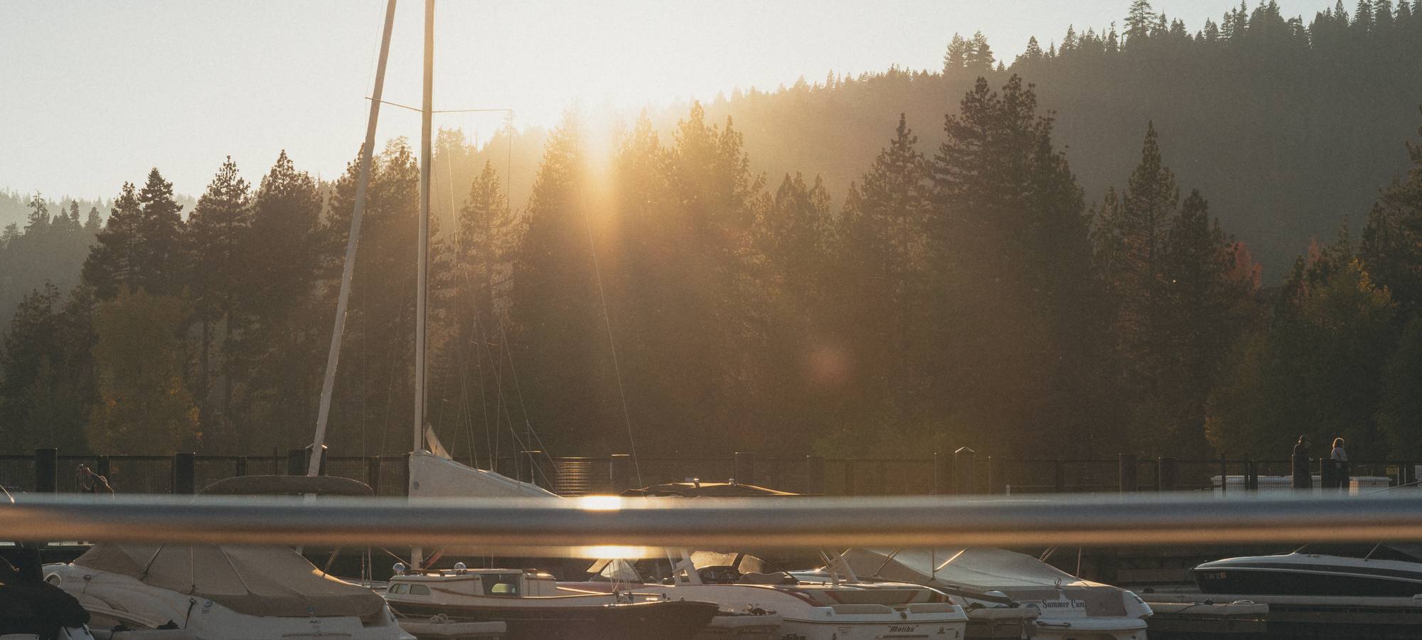Low sun through the pines over a Lake Tahoe marina, a sailboat mast and a dock rail in front.