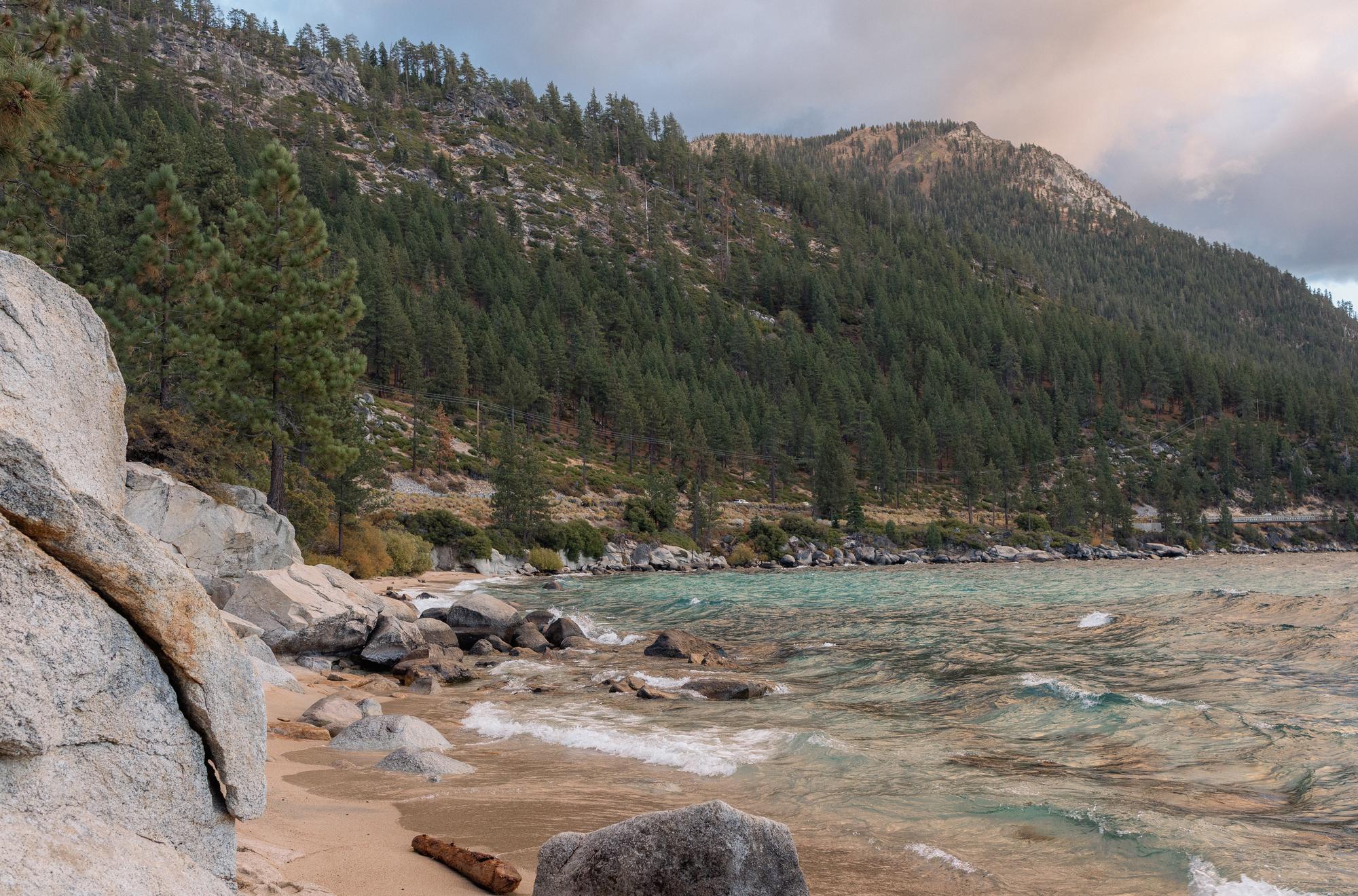 Waves washing into a small sand cove between granite boulders on Lake Tahoe's East Shore, pines on the slope above.
