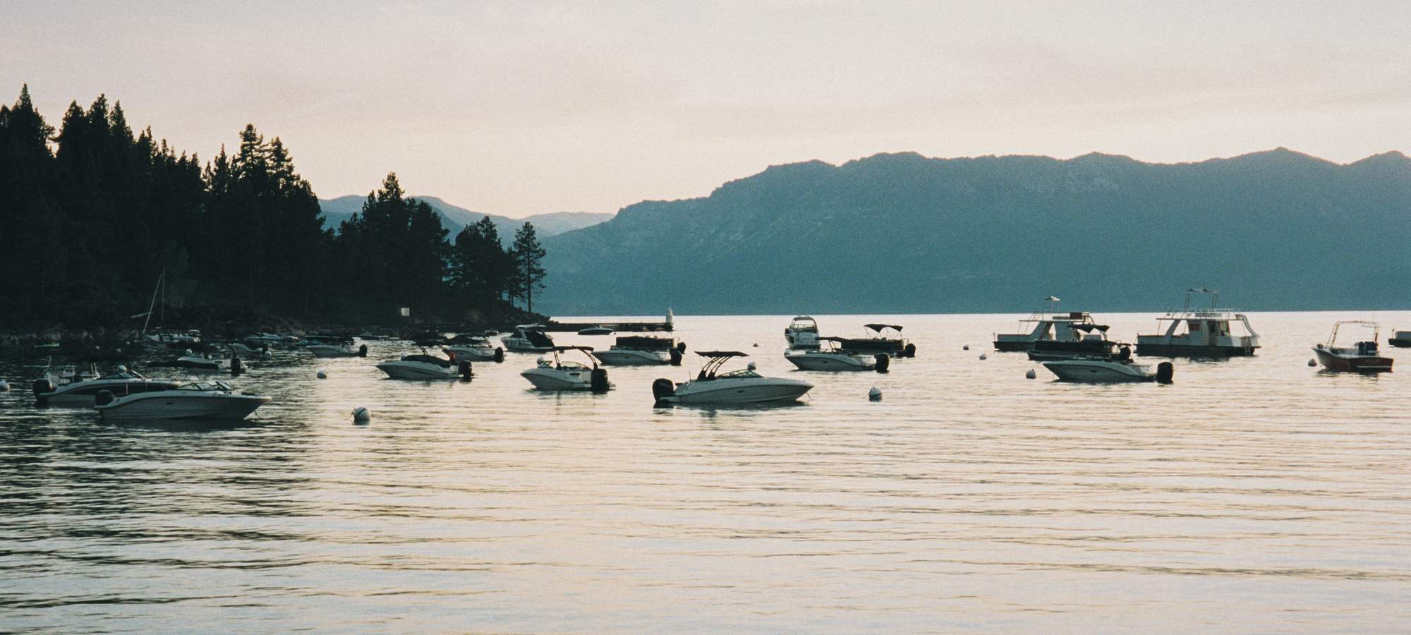 Boats at their moorings on calm Lake Tahoe, a pine-covered point and the far shore's mountains behind.