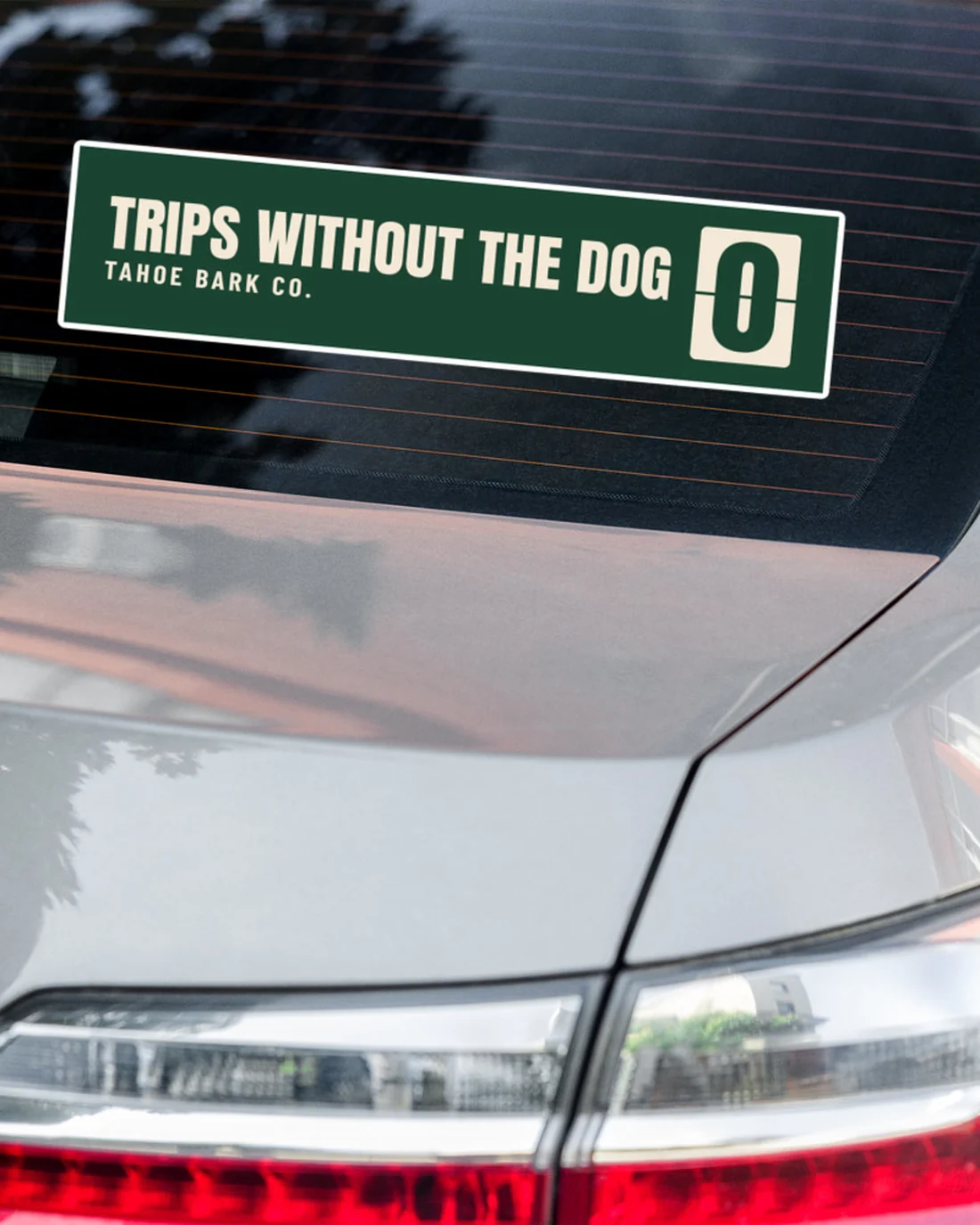 The Trips Without the Dog bumper sticker on a grey car's rear window, close up: TRIPS WITHOUT THE DOG and TAHOE BARK CO. in cream on pine beside the flip counter showing 0.